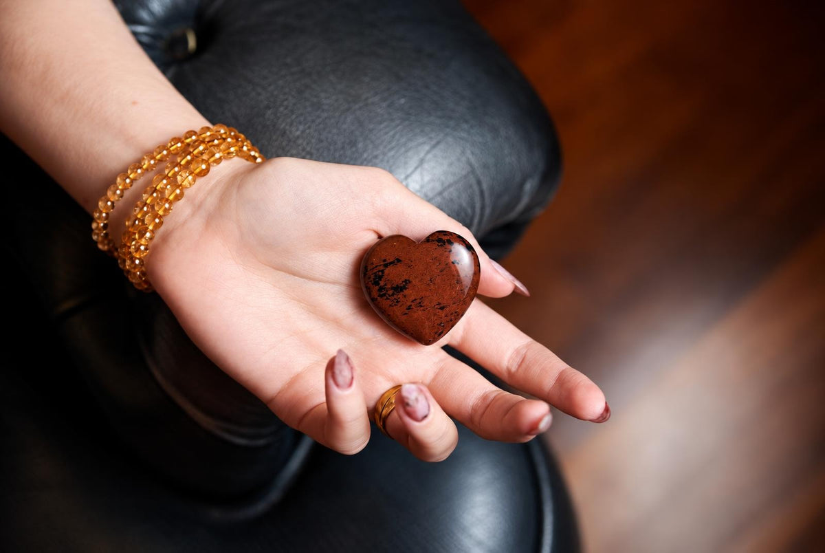 A hand holds a polished, heart-shaped stone with reddish-brown and black speckles against a dark leather surface.