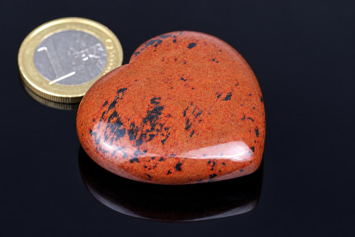 A polished reddish-brown heart-shaped stone with black patches rests beside a coin on a reflective black surface.