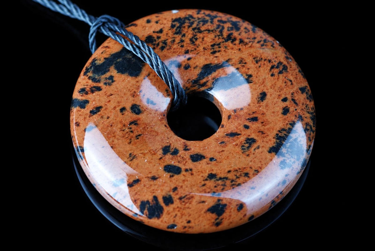 A polished, donut-shaped pendant with a glossy orange-brown surface speckled with black and white patches, threaded on a dark cord against a black background.