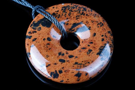 A polished, donut-shaped pendant with a glossy orange-brown surface speckled with black and white patches, threaded on a dark cord against a black background.