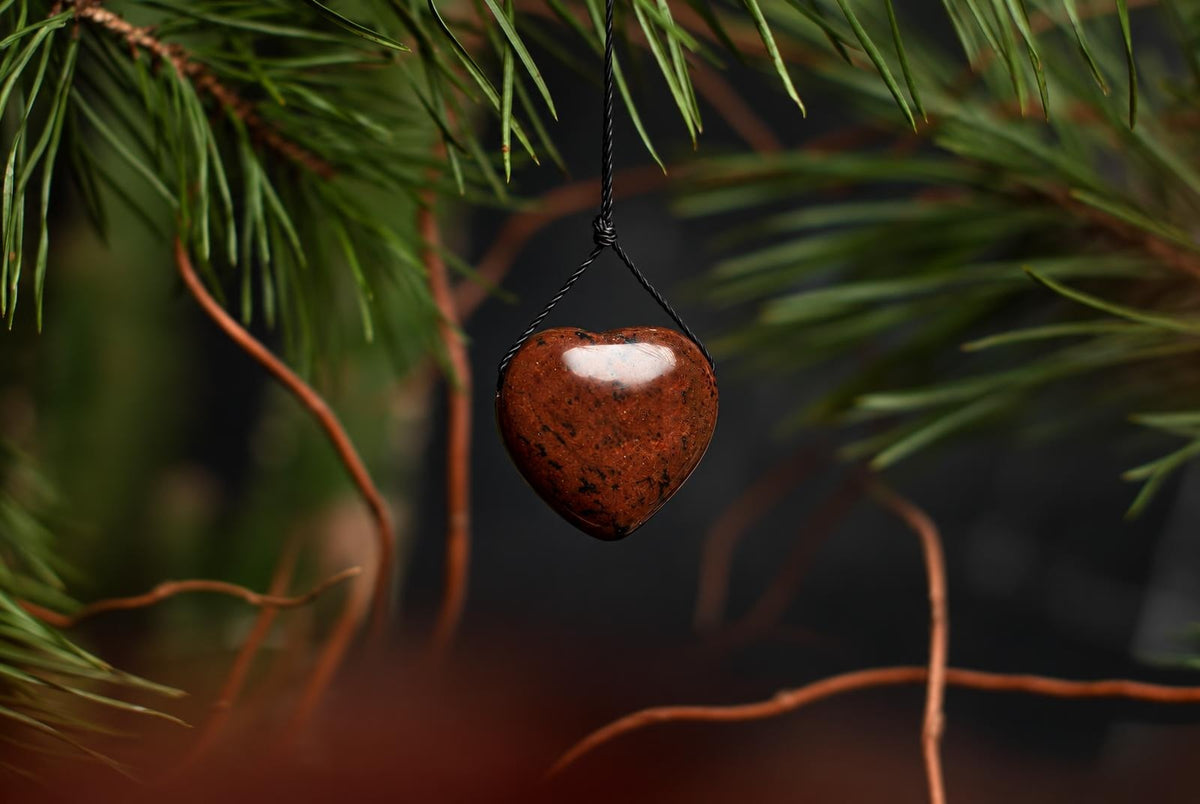 A reddish brown heart-shaped mahogany obsidian pendant with dark speckles hangs from a black cord among narrow green leaves.