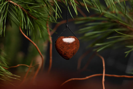 A reddish brown heart-shaped mahogany obsidian pendant with dark speckles hangs from a black cord among narrow green leaves.