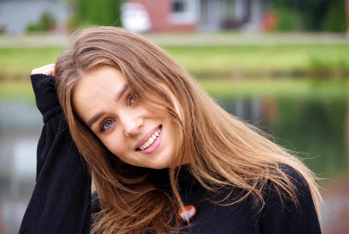 A smiling woman with long brown hair wearing a black top and a dark reddish-brown heart-shaped pendant, outdoors.