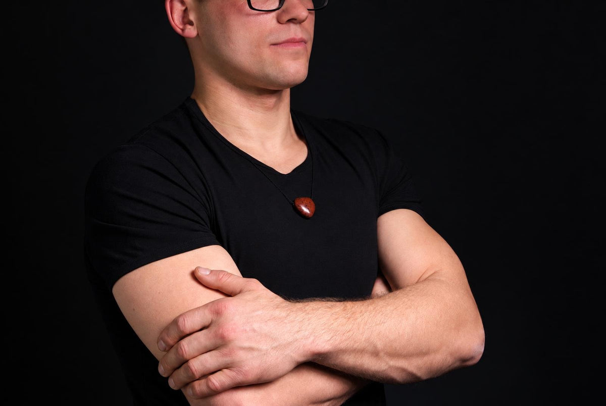 A man in a black shirt wears a necklace with a polished, reddish-brown heart-shaped pendant against a dark background.