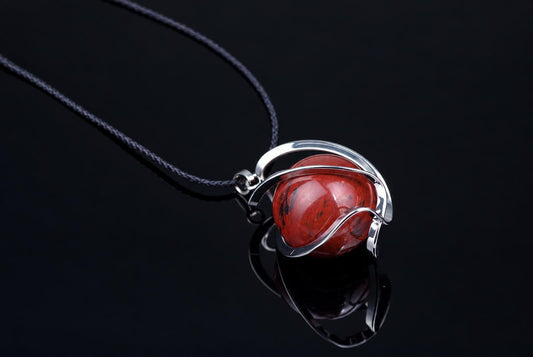 A silver-toned pendant with a red, speckled sphere rests on a dark surface.