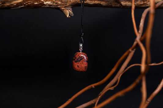 A polished red pendant with black speckles hangs from a black cord on a silver bail, suspended against a dark background near brown branches.