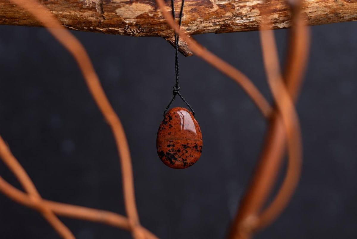 A polished, teardrop-shaped pendant with a reddish-brown base and black speckles hangs from a black cord against a dark background.