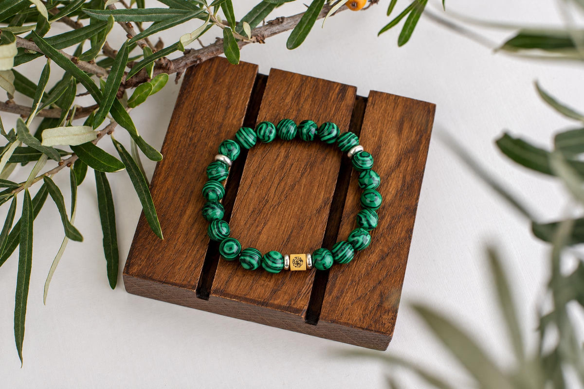 A bracelet with green patterned beads and a gold-toned square charm rests on a dark wooden surface, surrounded by foliage.