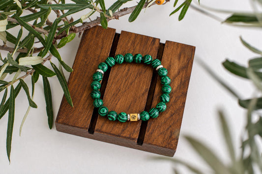A bracelet with green patterned beads and a gold-toned square charm rests on a dark wooden surface, surrounded by foliage.