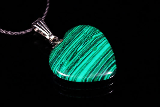 A heart-shaped pendant with green and black striped malachite on a dark surface, attached to a silver-toned bail and cord.