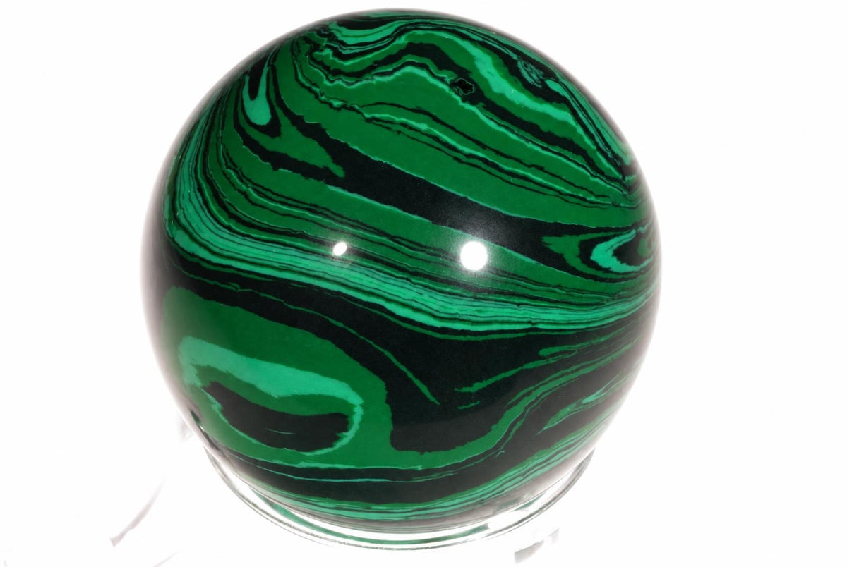 A polished sphere with swirling green and black patterns sits on a clear base against a white background.
