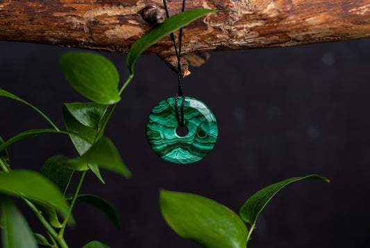 A polished green malachite pendant with a donut shape hangs from a black cord on a wooden branch, surrounded by green leaves.