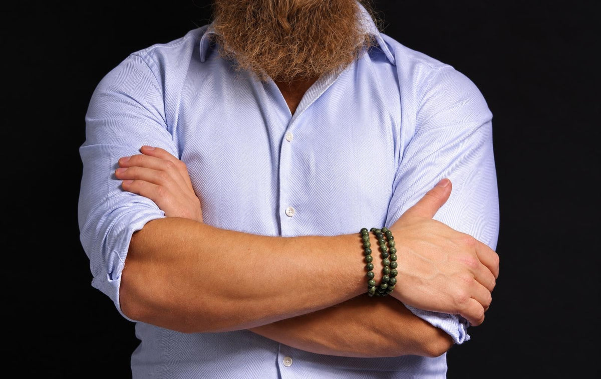 A person with a beard wears three dark green beaded bracelets on their wrist, arms crossed against a black background.