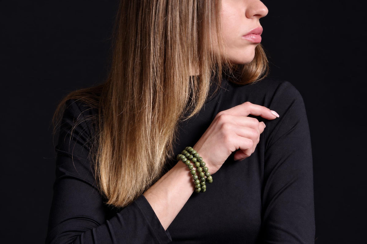 A woman with long blonde hair wears a dark top and a green beaded bracelet on her wrist against a black background.