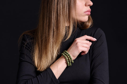 A woman with long blonde hair wears a dark top and a green beaded bracelet on her wrist against a black background.