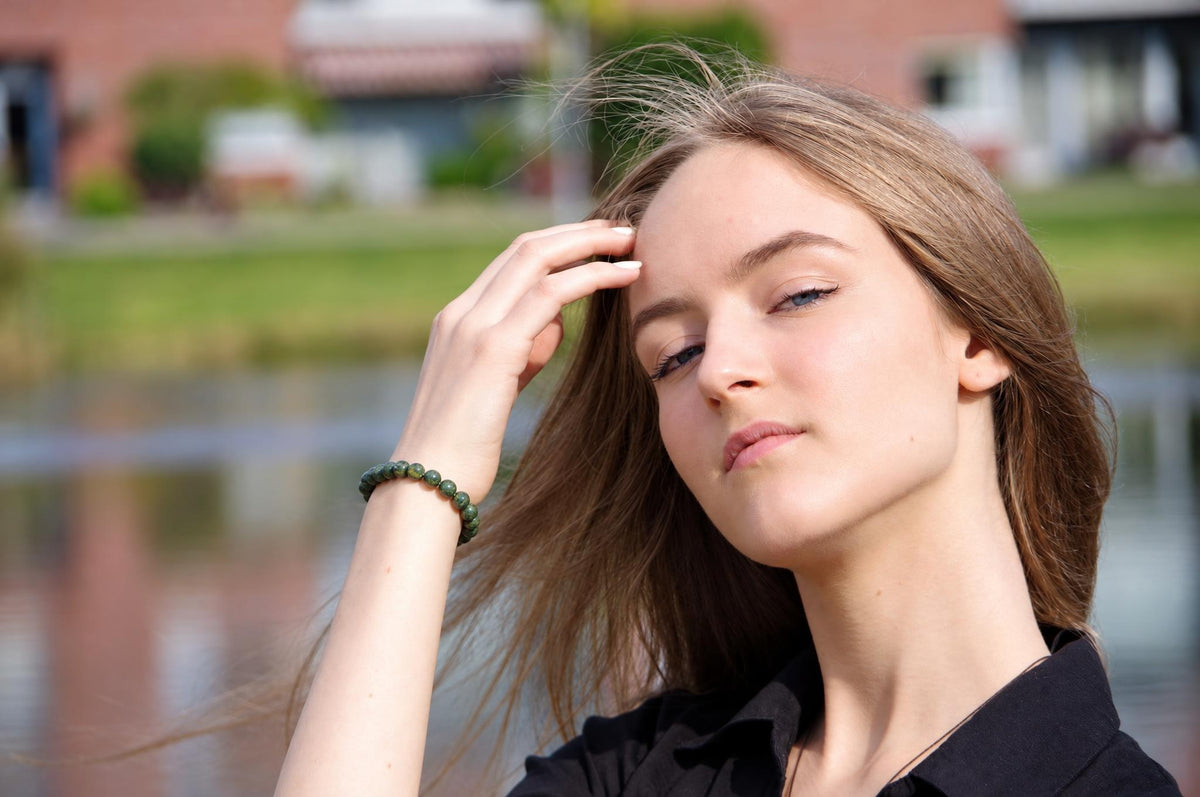 A woman wears a dark green beaded bracelet on her wrist, touching her hair outdoors near water.