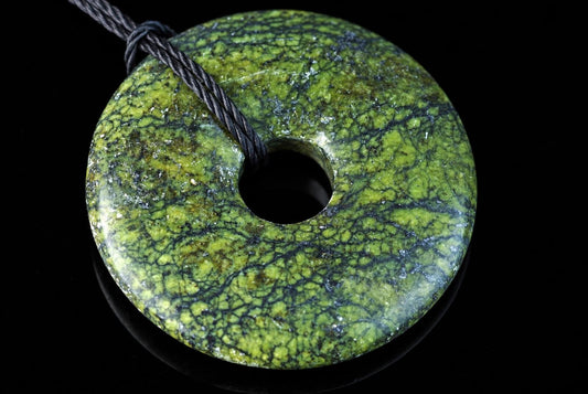 A green and black patterned donut-shaped pendant with a central hole, threaded on a dark cord against a black background.