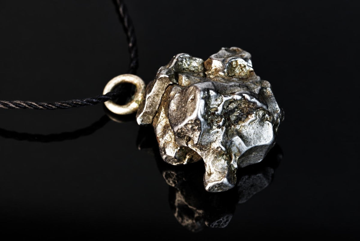 A silver-toned, irregularly shaped pendant with a rough texture hangs from a black cord against a dark, reflective surface.