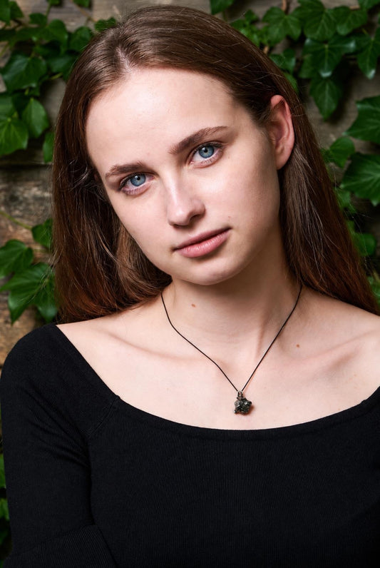 A woman wears a black top and a necklace with a dark, irregular pendant on a thin cord against a leafy background.