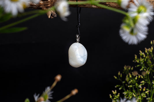 A white pendant with faint blue swirls hangs from a black cord against a dark background, framed by flowers.