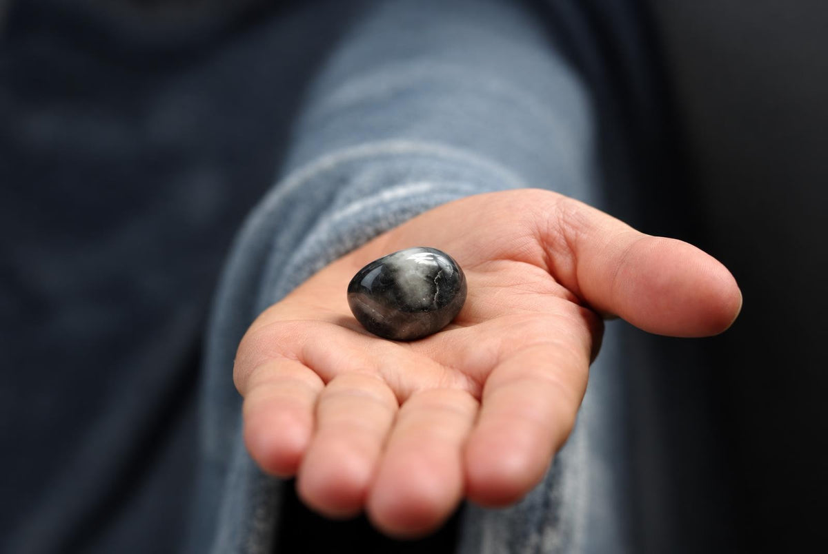 A hand holds a smooth, polished stone with dark and white cloudy patterns on a dark surface.