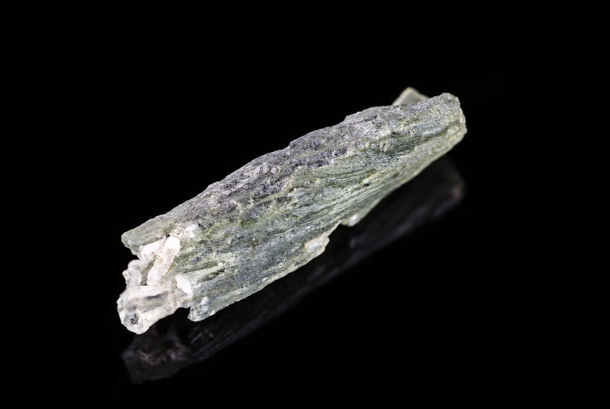 A green, elongated mineral specimen with a rough texture and white crystalline inclusions rests on a dark surface.