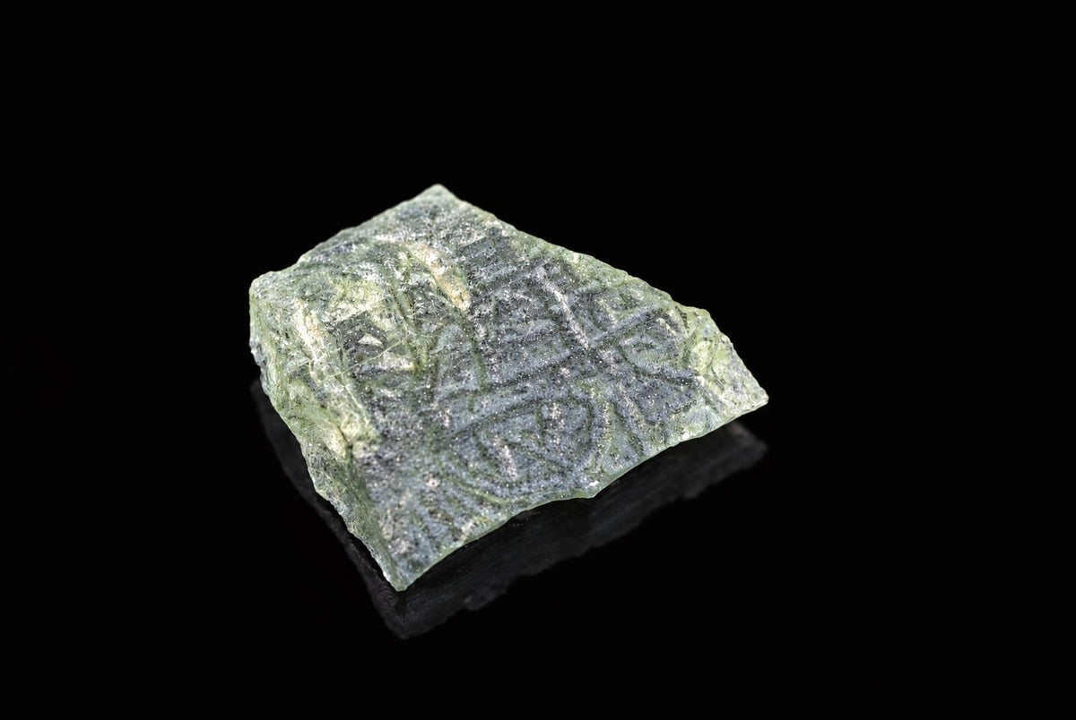 A green, translucent Moldavite stone with a rough, angular shape and internal patterns rests on a dark surface.