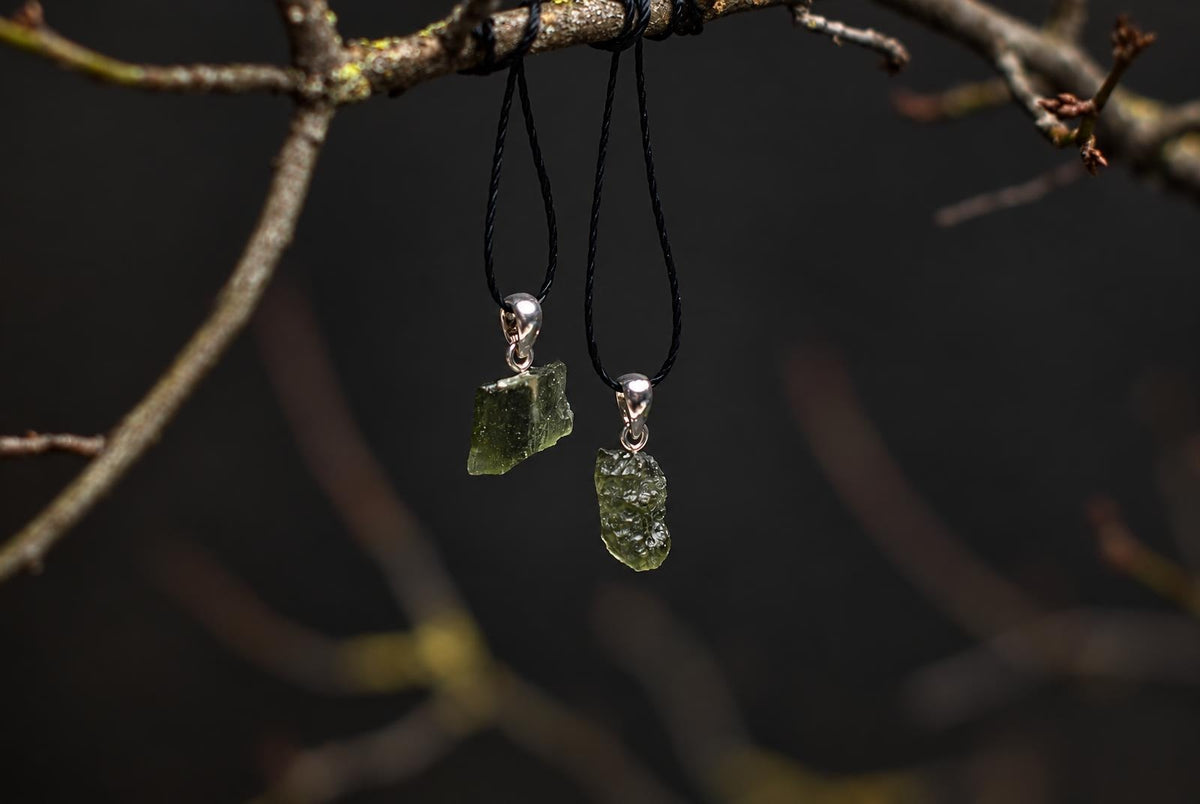 Two green, irregular glass pendants hang from black cords on a dark branch against a blurred background.