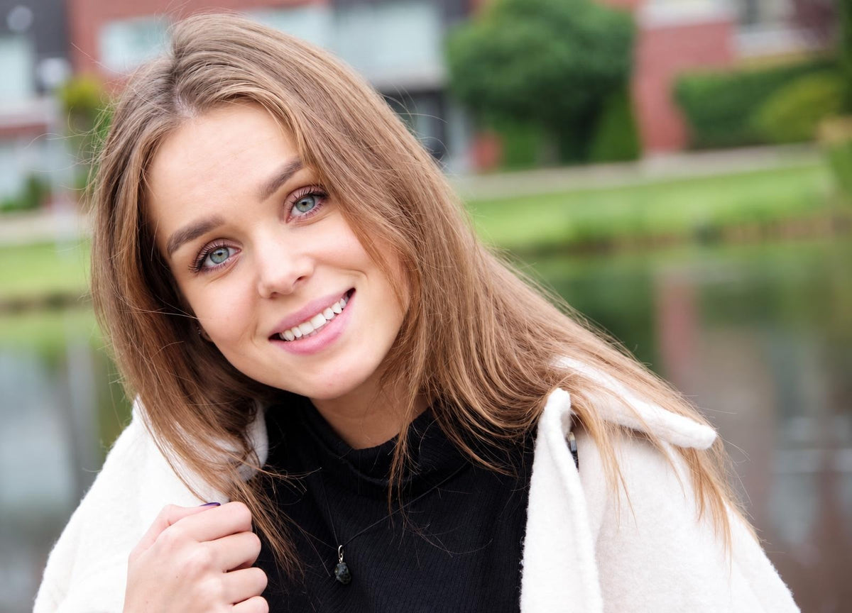 A smiling woman with long brown hair wears a white coat over a black top and a dark pendant necklace outdoors.