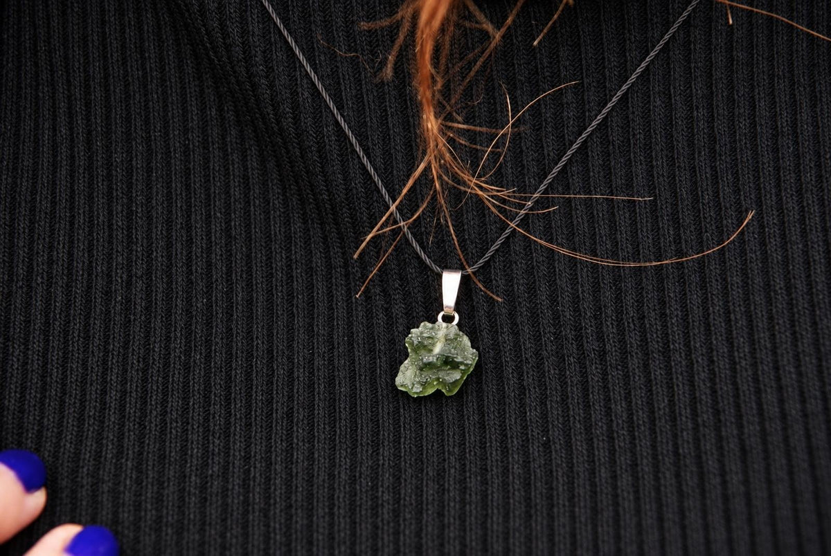 A green, irregularly shaped pendant hangs from a dark cord on a black ribbed fabric surface.
