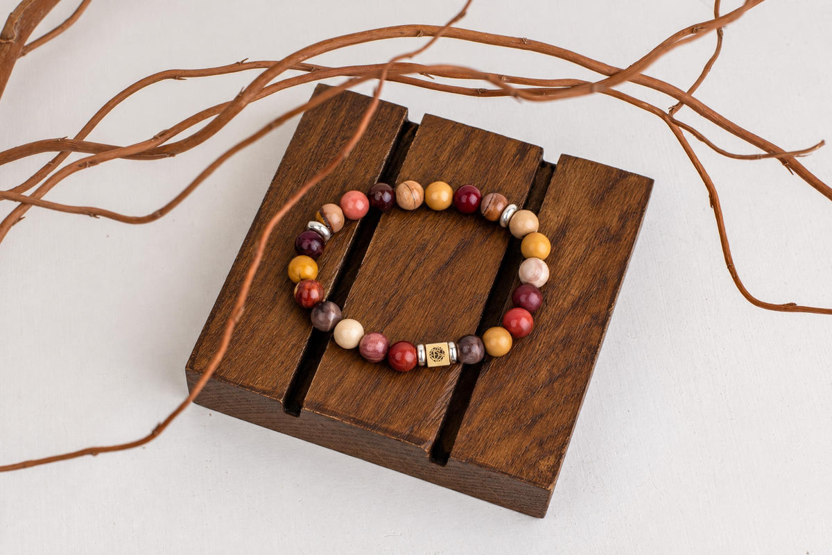 A beaded bracelet with round, multi-coloured stones and a square silver-toned charm rests on a dark wooden stand.