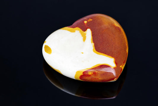 A polished heart-shaped stone with white, red and yellow patterns on a dark reflective surface.