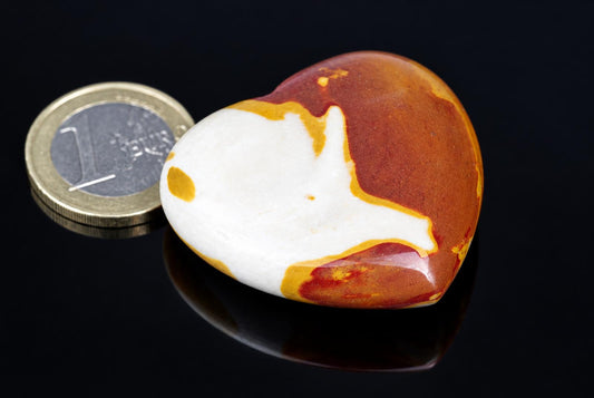A polished heart-shaped stone with white, orange and red patterns sits on a dark surface next to a one-euro coin.