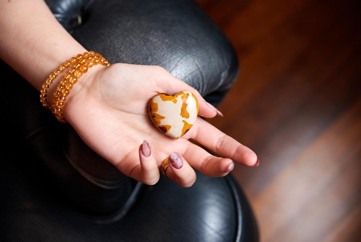 A hand holds a heart-shaped stone with cream and brown mottled patterns, resting on a dark leather surface.