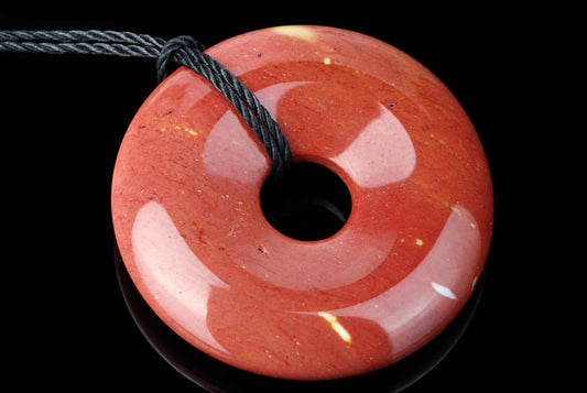 A polished, reddish-orange donut-shaped pendant with a central hole and black cord against a dark surface.