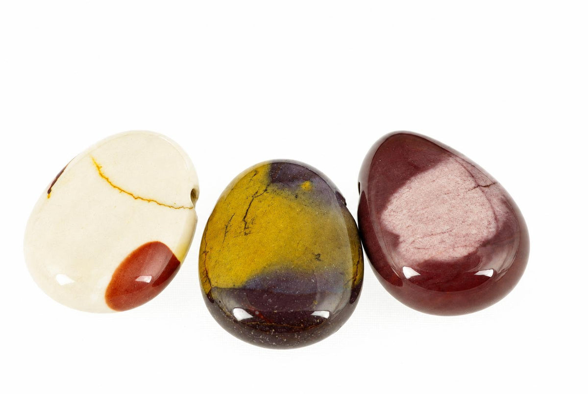 Three polished, teardrop-shaped beads on a white surface. The left bead is cream with red and yellow markings; the middle one is yellow and purple; the right one is dark red with a pale pink patch.