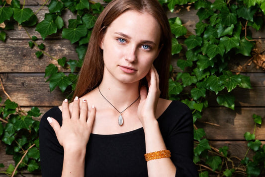 A woman in a black top wears a pale oval pendant on a thin black cord in front of a wooden wall covered with green leaves.