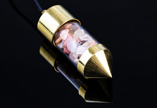 A brass pendulum with a pointed tip and cylindrical body, filled with pink and white stones, on a dark reflective surface.