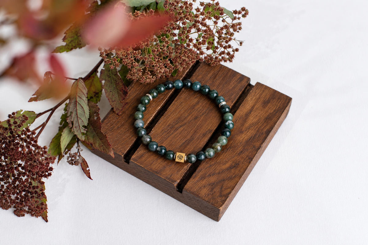 A bracelet of green round beads with a small square metal bead rests on wooden slats beside reddish leaves and dried flower clusters.