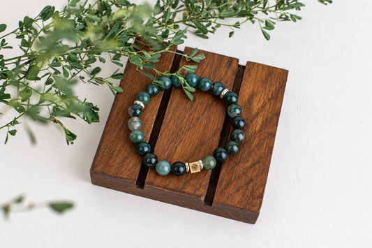 A beaded bracelet with green and dark beads, a gold-toned square charm, and silver spacers rests on a wooden stand beside green leaves.