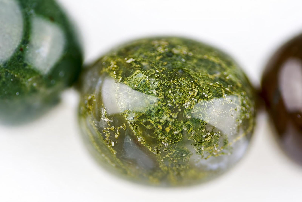 A close-up of a round, polished bead with green moss-like inclusions and white swirls on a pale surface.