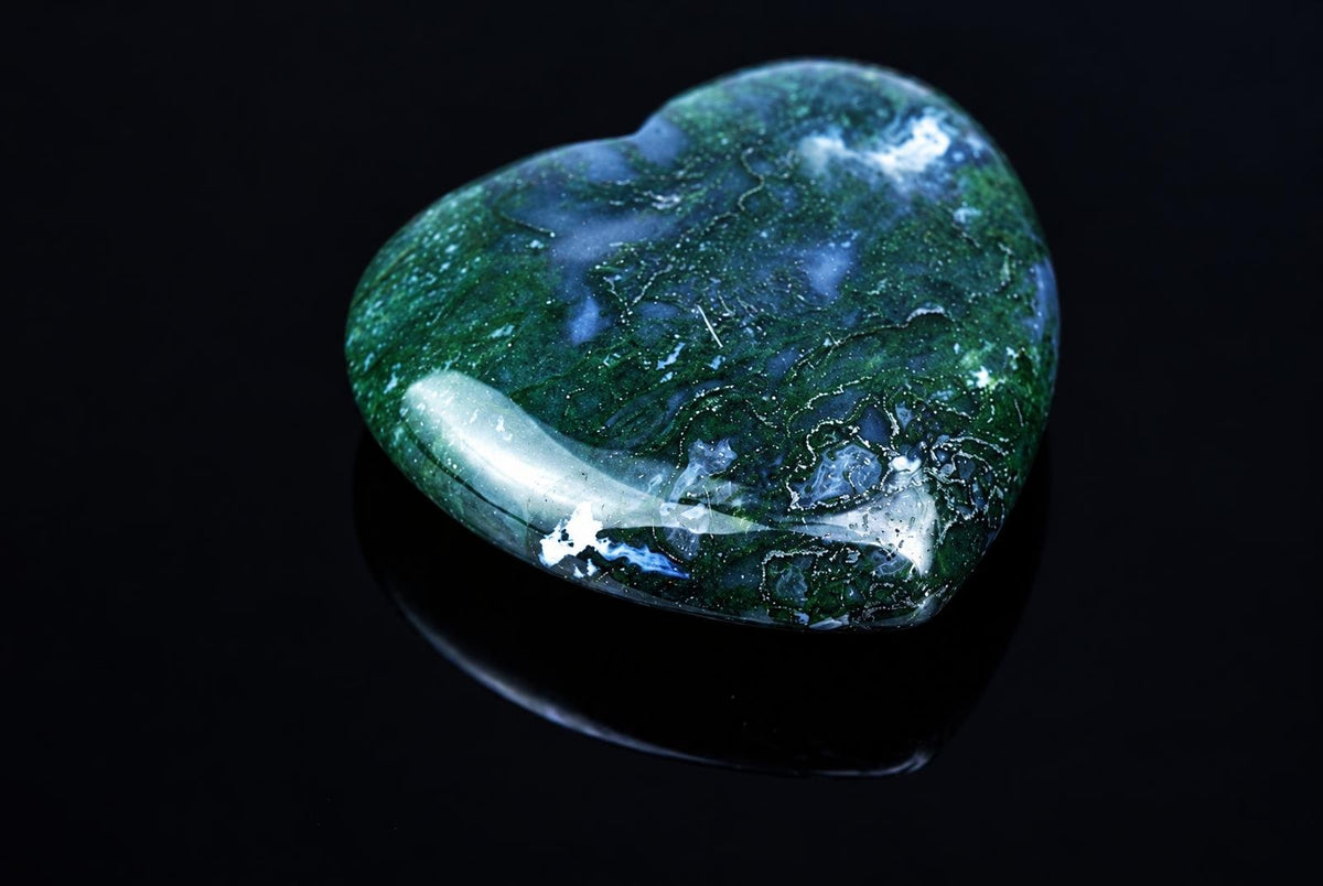 A polished heart-shaped stone with green and white swirling patterns on a dark reflective surface.