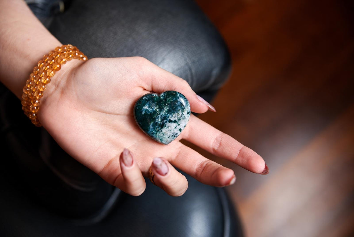 A hand holds a polished heart-shaped stone with green and white patterns on a dark surface.
