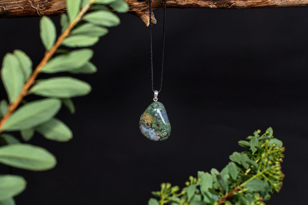 A teardrop-shaped pendant with green, blue and brown patterns hangs from a black cord on a dark surface.