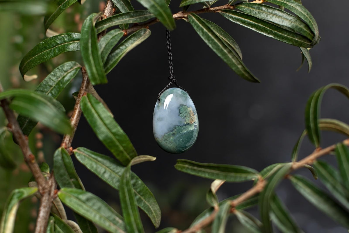 A polished, oval-shaped pendant with light blue and green moss agate patterns hangs from a black cord against dark foliage.