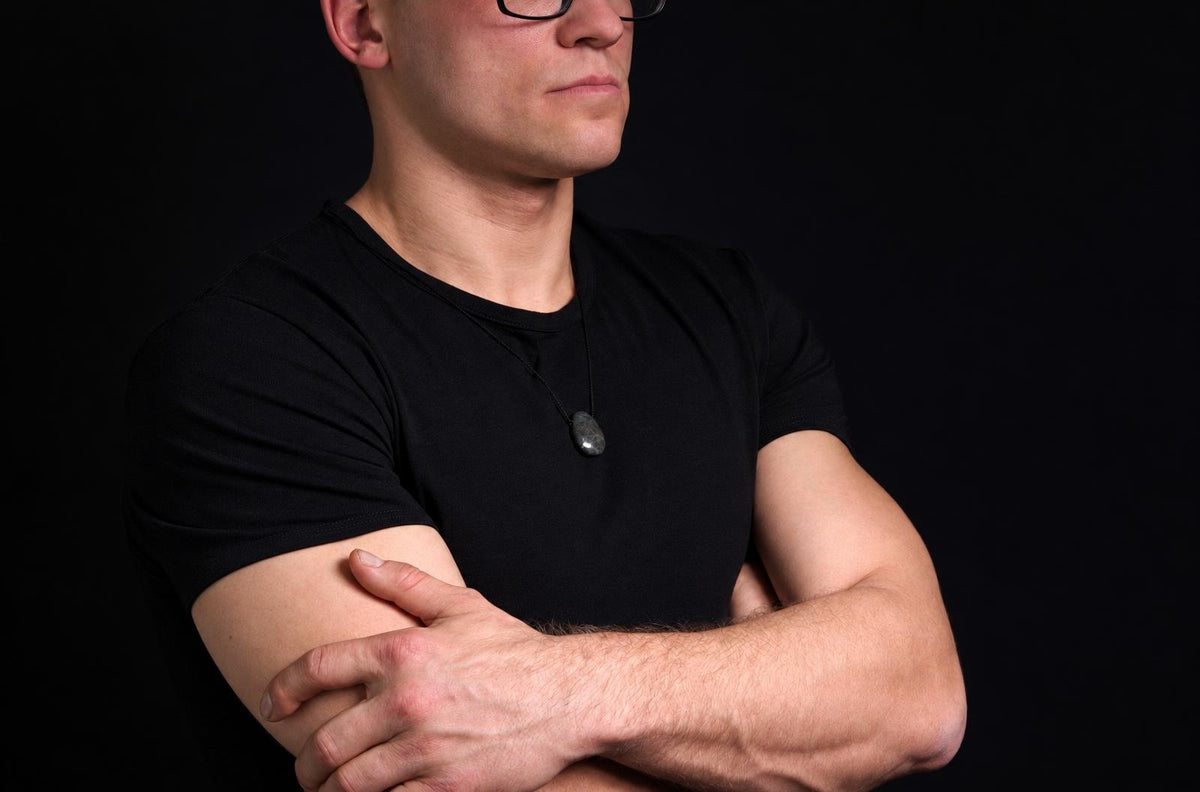 A man in a black t-shirt wears a dark, smooth oval pendant on a thin cord against a dark background.