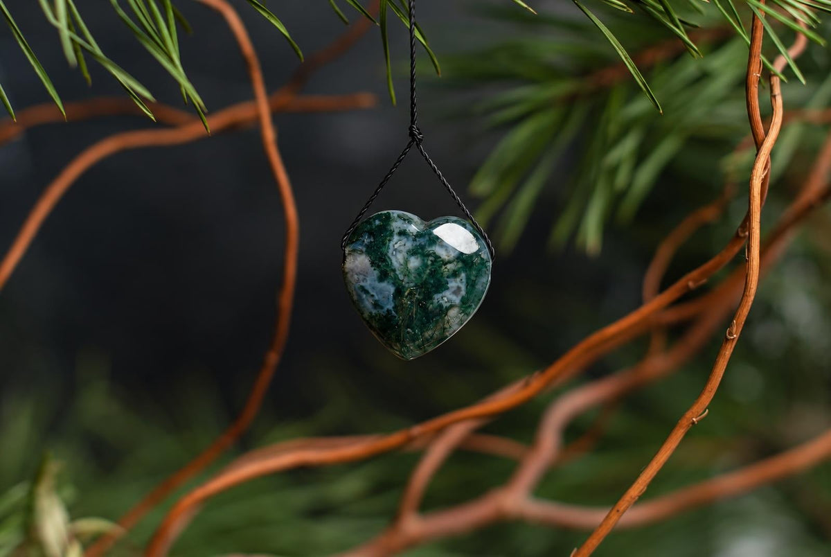 A polished heart-shaped pendant with green and white mottled patterns hangs from a black cord against blurred branches.