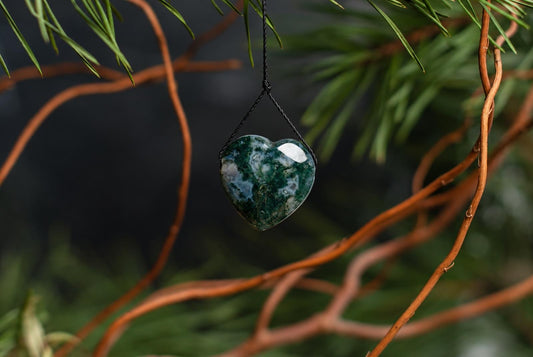 A polished heart-shaped pendant with green and white mottled patterns hangs from a black cord against blurred branches.