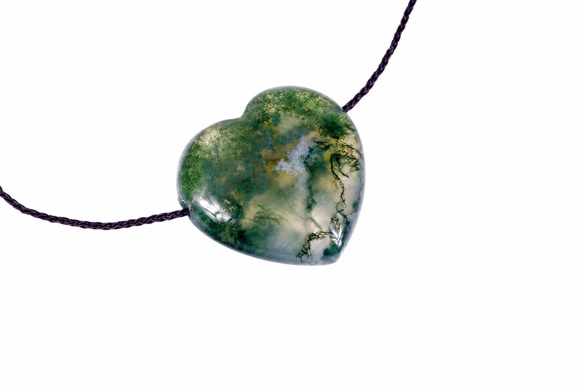 A heart-shaped moss agate pendant with green, white and yellow inclusions on a dark cord against a pale surface.