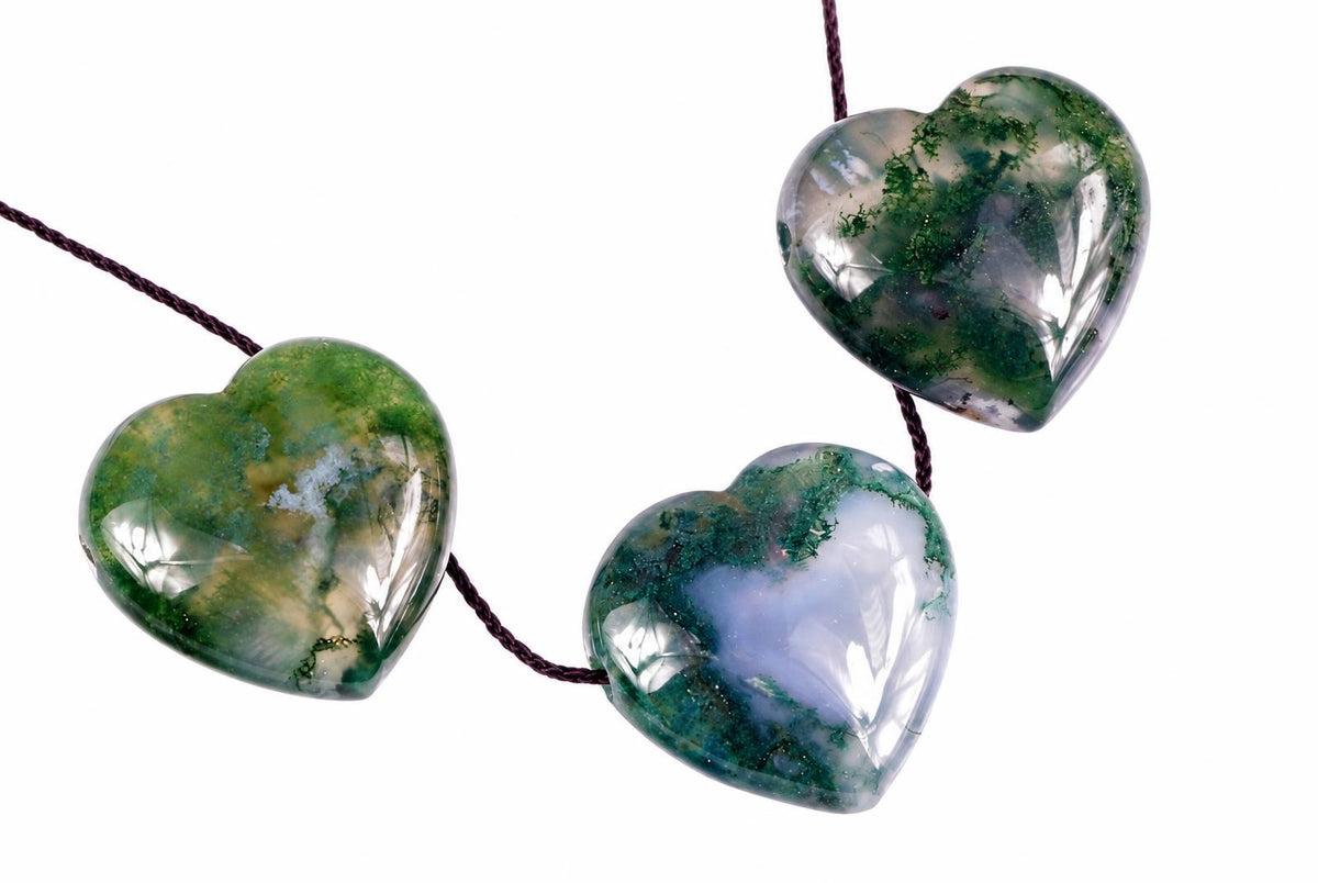 Three heart-shaped moss agate pendants with green and white patterns hang from dark cords against a white background.